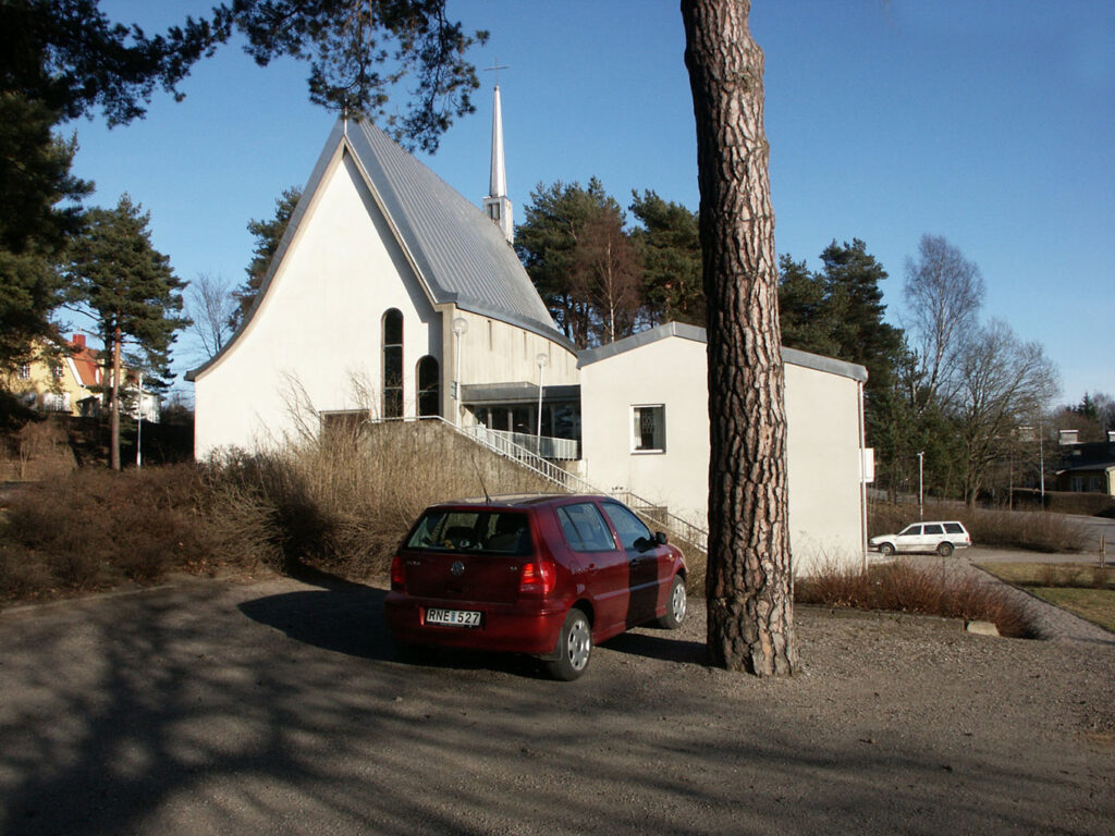White church with steep roof and tall spire surrounded by trees, with parked cars in a gravel lot under a clear blue sky.