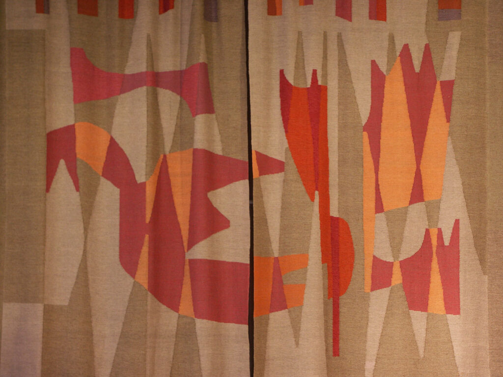 Abstract woven curtain with geometric shapes in muted beige tones and bold red and orange forms creating a dynamic mid-century pattern.