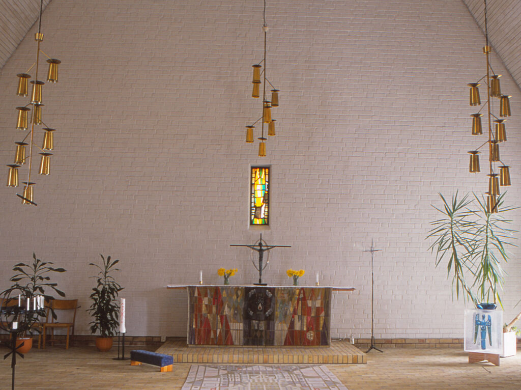 Modern church interior with brick walls, hanging brass lights, a colorful altar cloth, a central sculpture, plants, and a small stained-glass window.