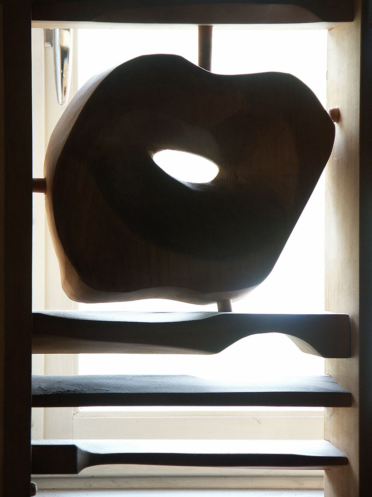Carved wooden abstract sculpture with a rounded organic form and a central hole, displayed against bright window light on wooden shelves.