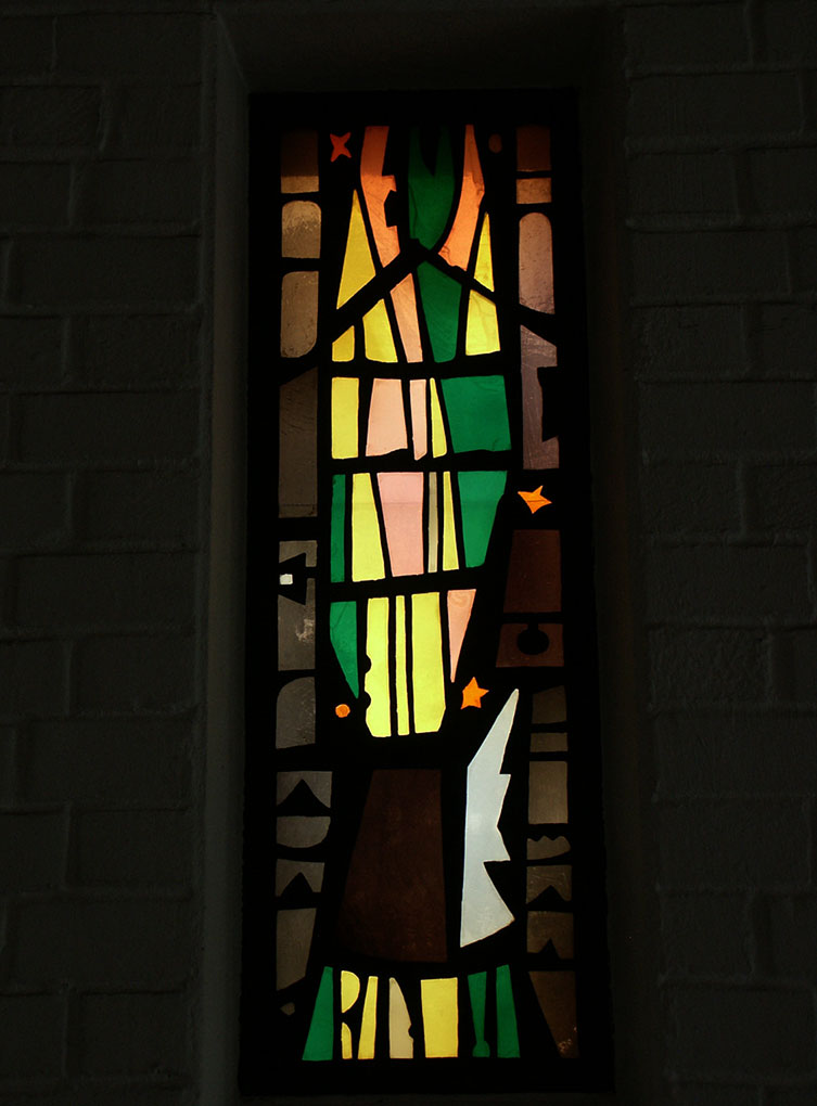 Modern church interior with brick walls, hanging brass lights, a colorful altar cloth, a central sculpture, plants, and a small stained-glass window.