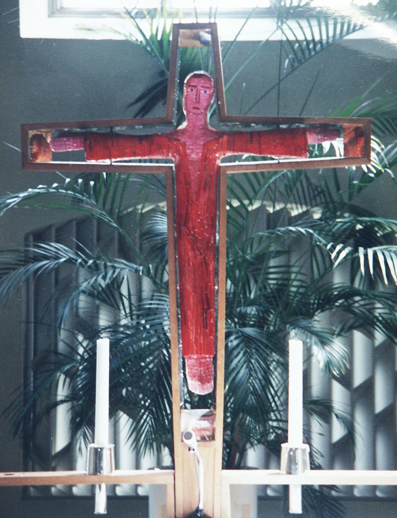 Wooden cross with a stylized red painted figure at its center, flanked by two white candles, set before tall green indoor plants.