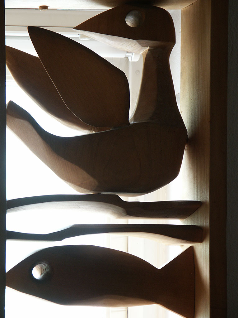 Carved wooden bird with outstretched wings above a stylized fish, both set in a window display with strong backlighting.