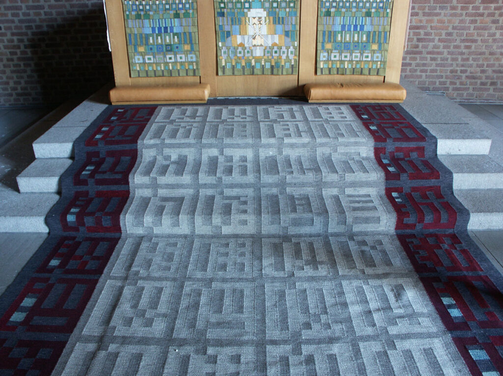 Patterned gray carpet with geometric designs running up church steps, bordered by maroon motifs, leading to a colorful mosaic altarpiece.