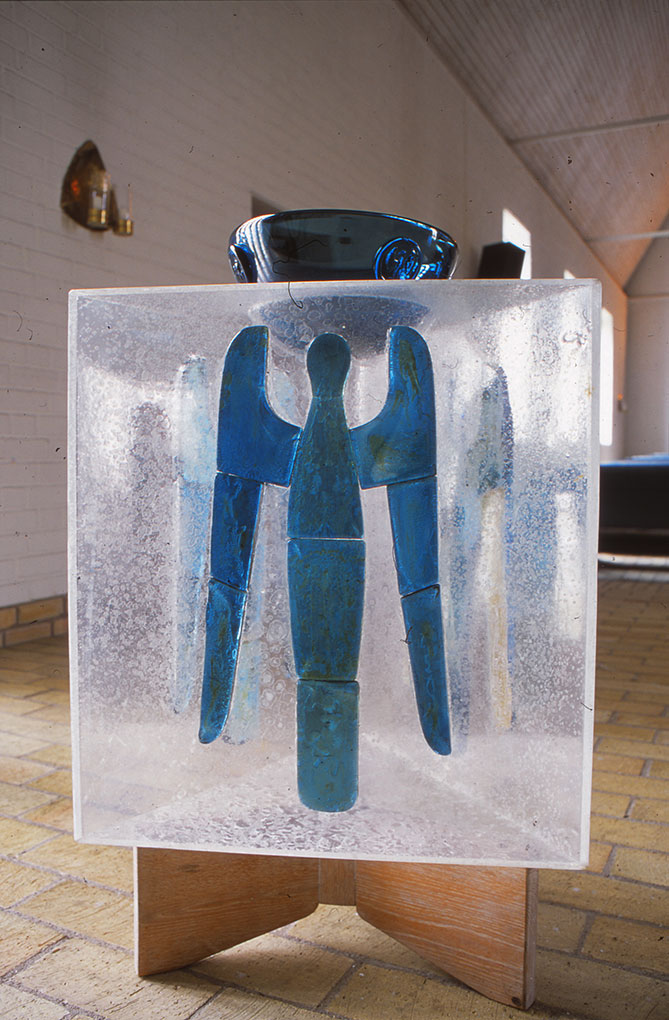 Blue angel figure encased in a translucent block on a wooden stand, with a dark glass bowl on top, set in a bright modern church interior.