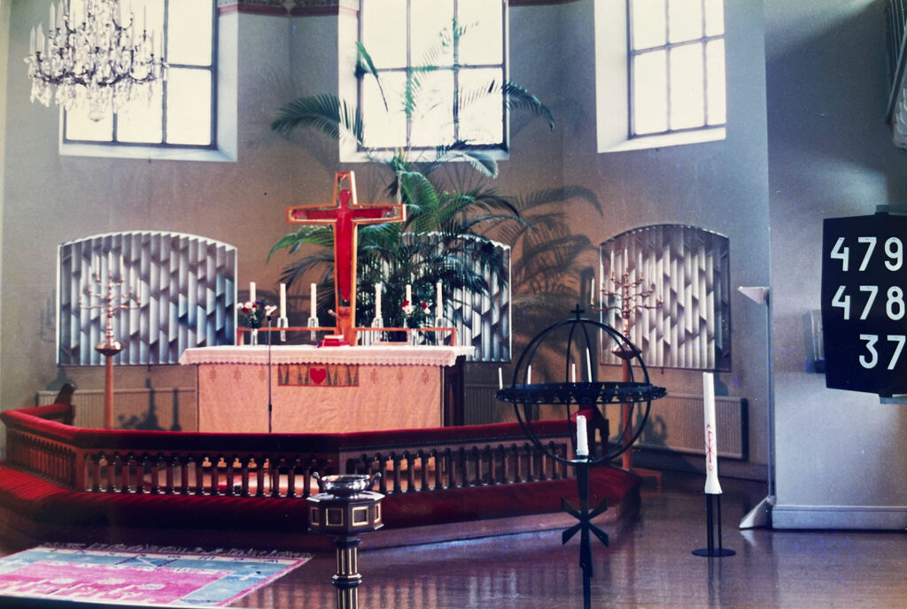 Church altar with a red cross, candles, plants, chandeliers, patterned wall panels, and hymn numbers displayed on a black board.
