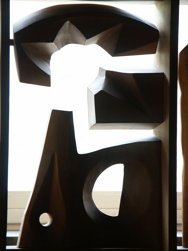 Tall carved wooden sculpture with angular geometric shapes and circular cutouts, silhouetted against a bright window.