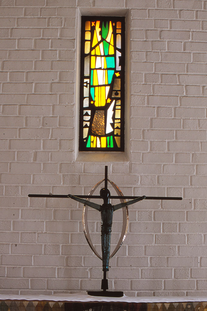 Tall stained-glass window above a modern metal crucifix with a circular halo, set against a white brick wall in a minimalist church interior.