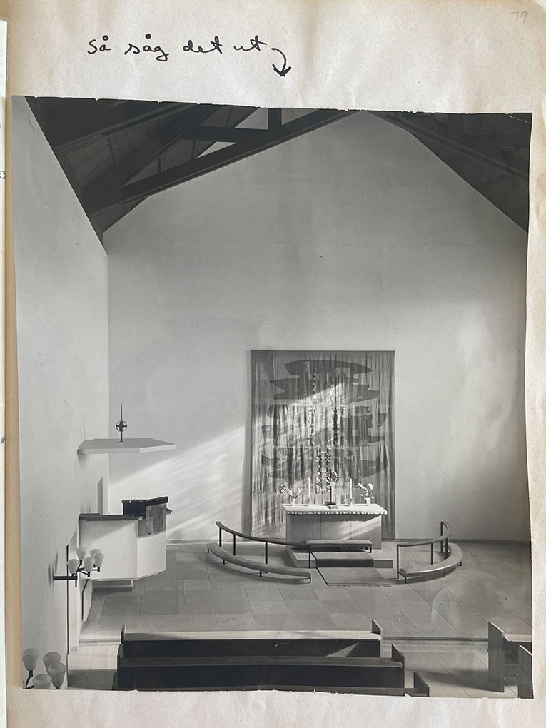 Black-and-white photo of a church interior with simple altar, curved rail, wall hanging, wooden beams, and handwritten note at top.