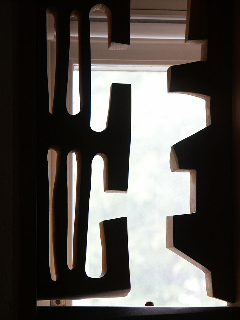 Vertical wooden sculpture of narrow elongated cut-outs and angular forms, shown in silhouette before a softly lit window.
