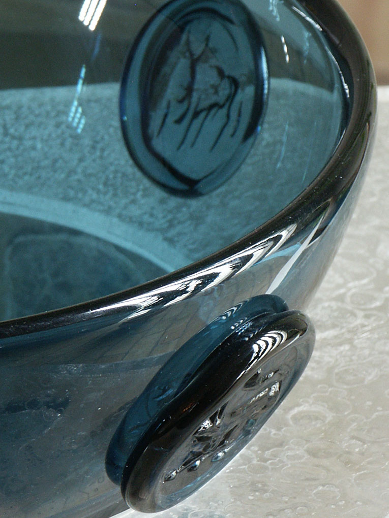 Close-up of a blue glass bowl with raised circular medallions on the sides, showing textured, etched designs and light reflections on the surface.