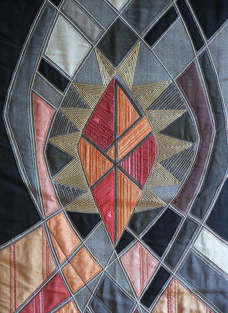 Colourful embroidered textile with geometric shapes forming a radiant central motif in gold, red, and orange against dark stitched panels.