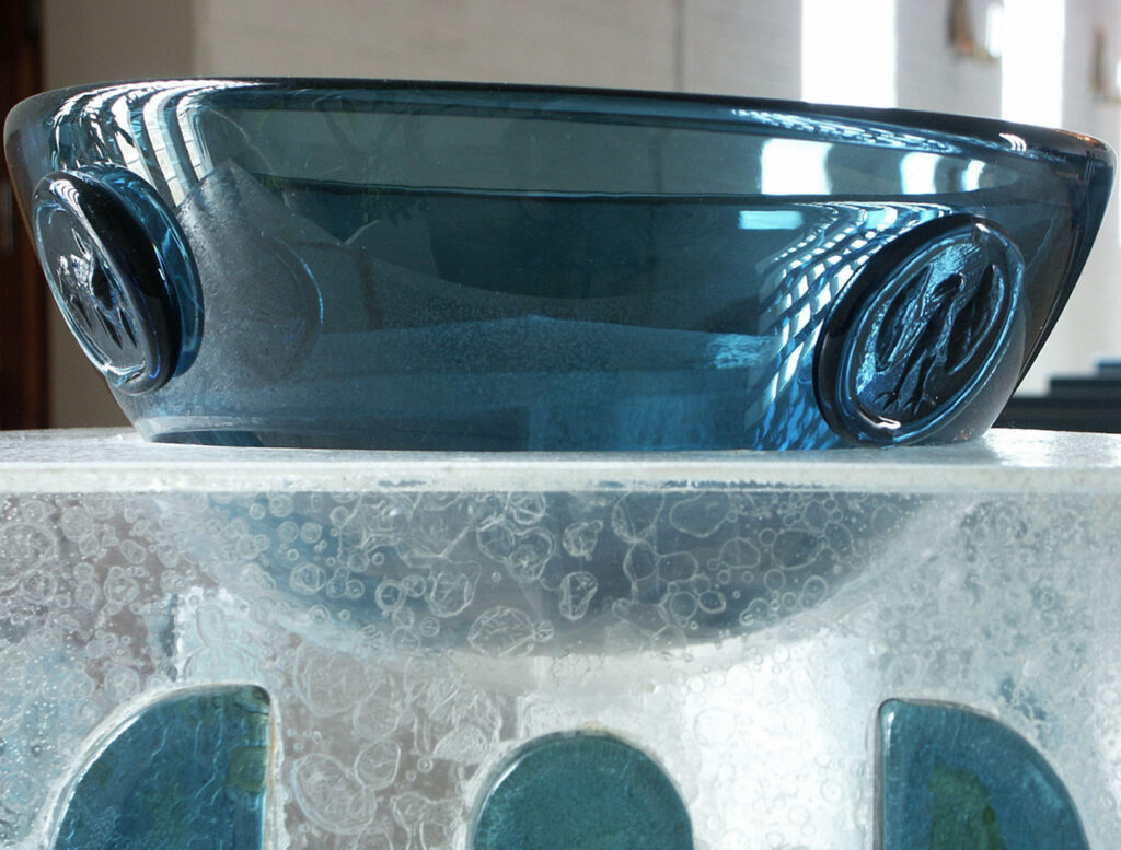 Blue glass bowl with embossed medallions sits atop a textured, frosted glass pedestal featuring blue inlaid shapes, lit by soft indoor light.