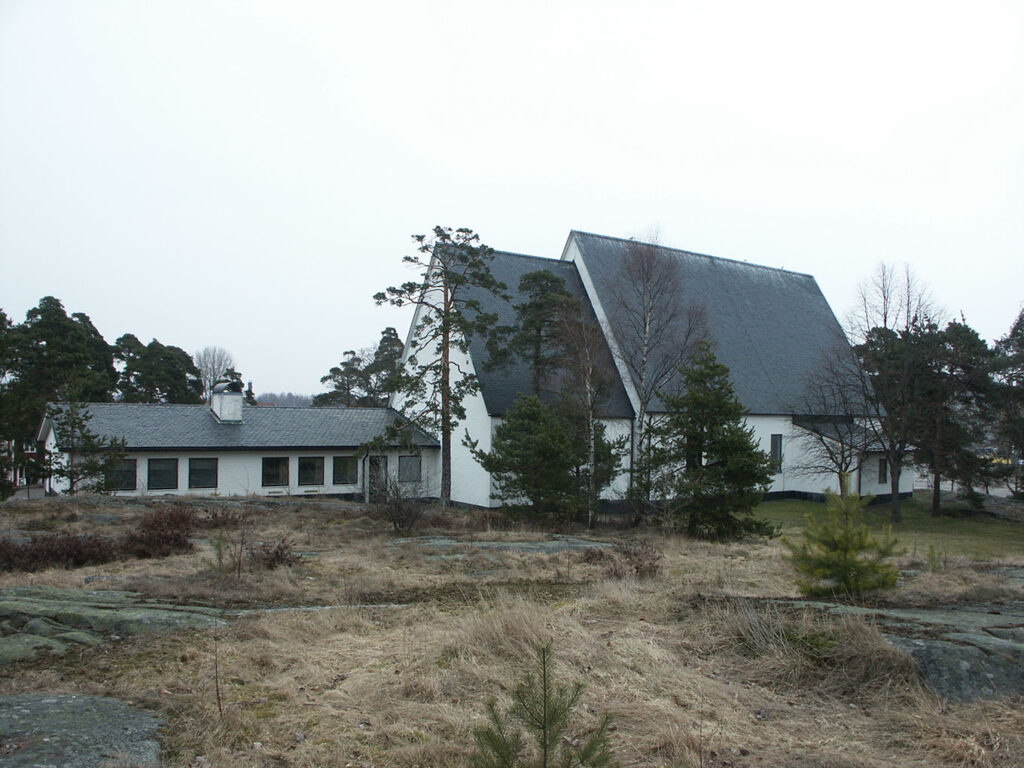 White modern church building with steep dark roofs surrounded by sparse trees and rocky, dry grassland under an overcast sky.