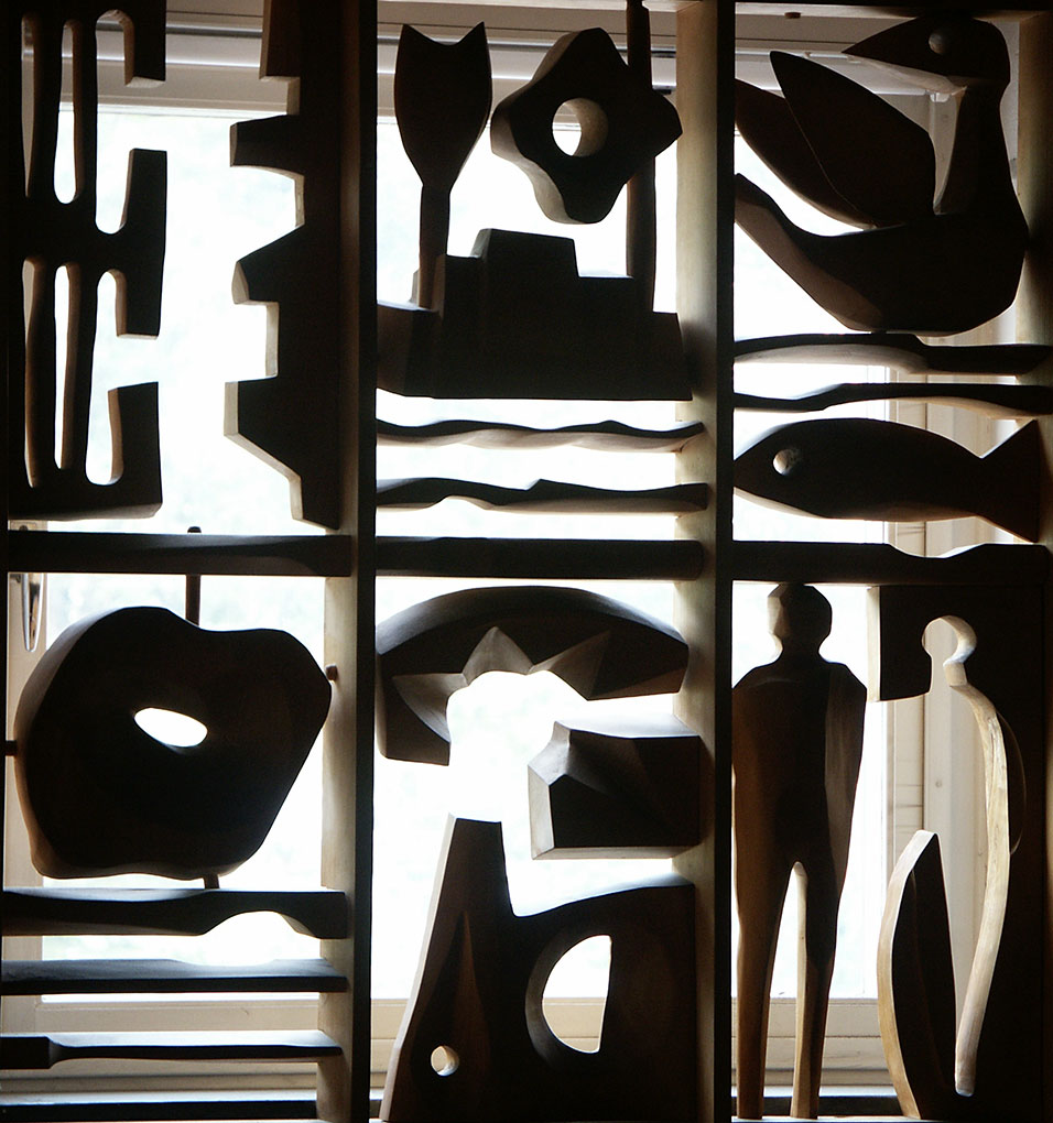 Panel of carved wooden sculptures with abstract shapes, birds, fish and figures arranged in a grid against bright window light.