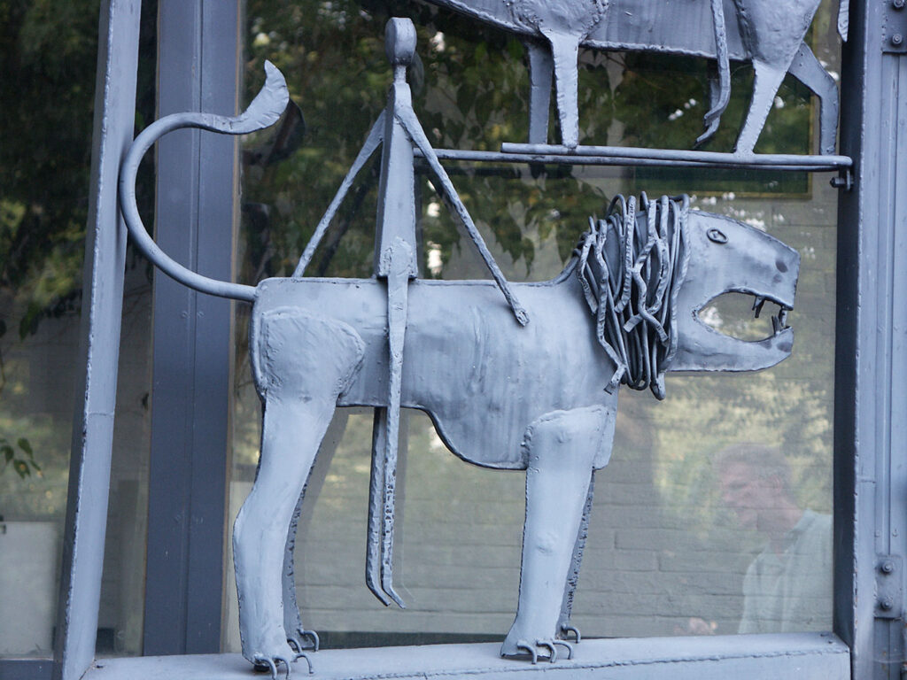 Close-up of a welded metal lion sculpture with an open mouth and textured mane, mounted on a glass wall outdoors.