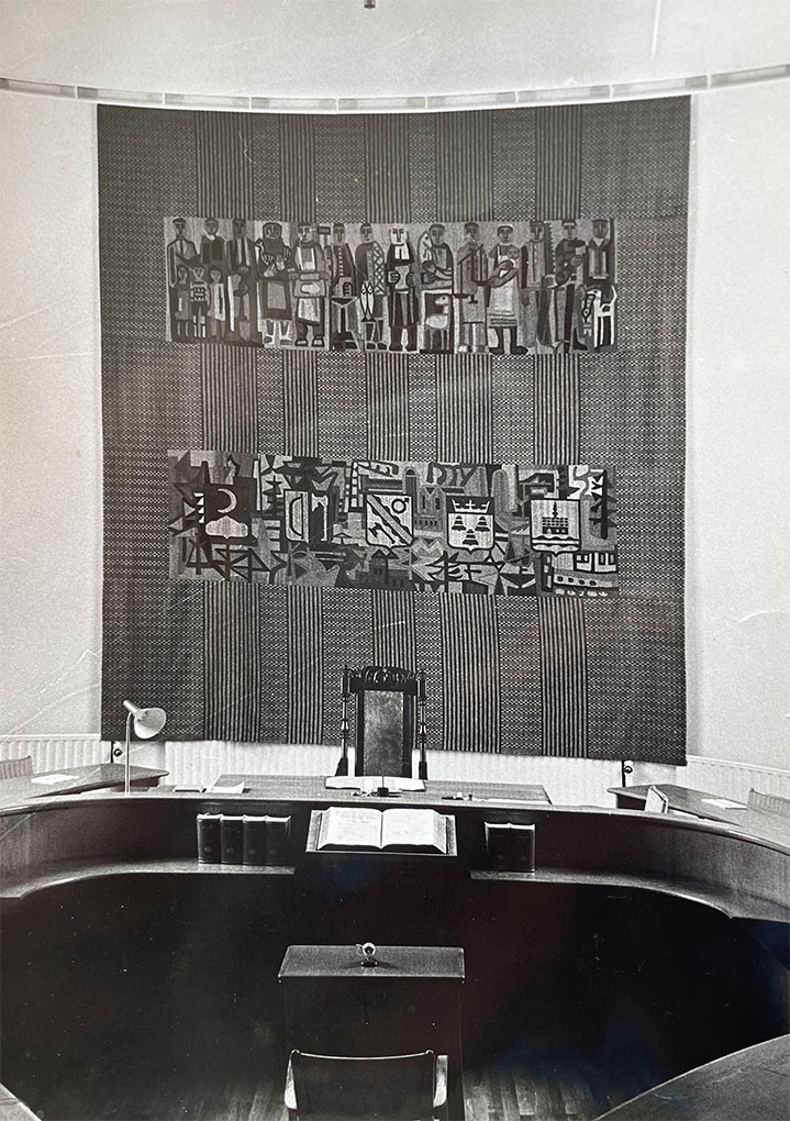 Black-and-white photo of a courtroom interior featuring a large tapestry with rows of figures and abstract motifs hanging behind the judge’s seat.