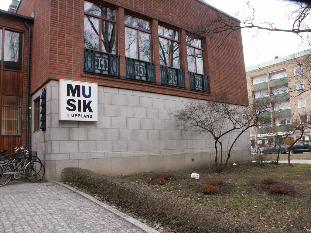 Street view of brick music building with abstract metal balcony panels, sign reading ‘Musik i Uppland,’ bikes and bare trees outside.