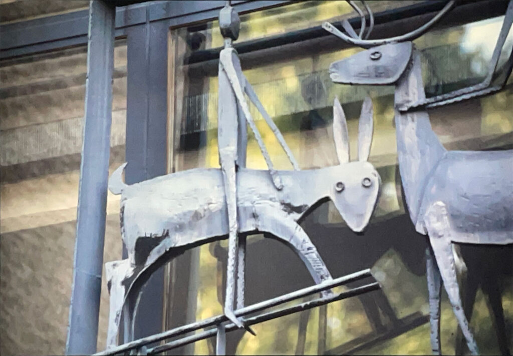 Detail of a metal sculpture showing a stylized figure riding a goat with long ears, mounted on a window frame.