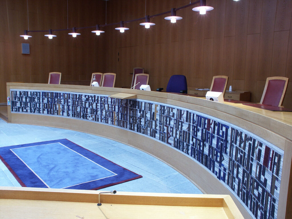 Curved courtroom bench displaying a long woven panel of abstract letter-forms beneath judges’ seats.