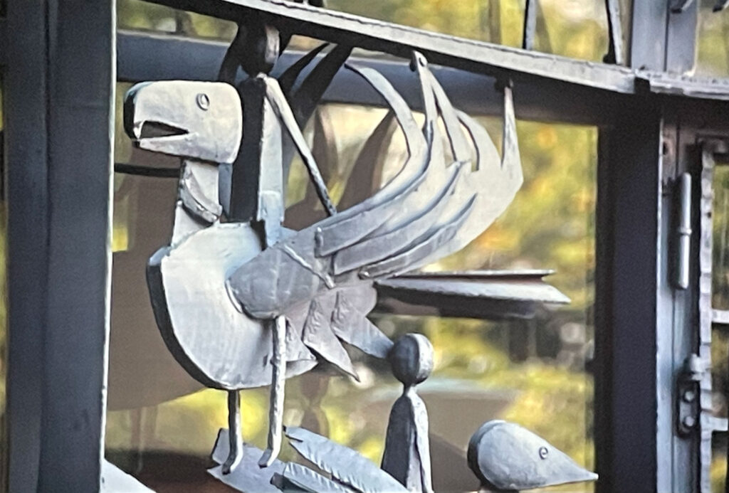 Close-up of a metal bird sculpture with layered wings and a perched figure beneath, attached to a building façade.