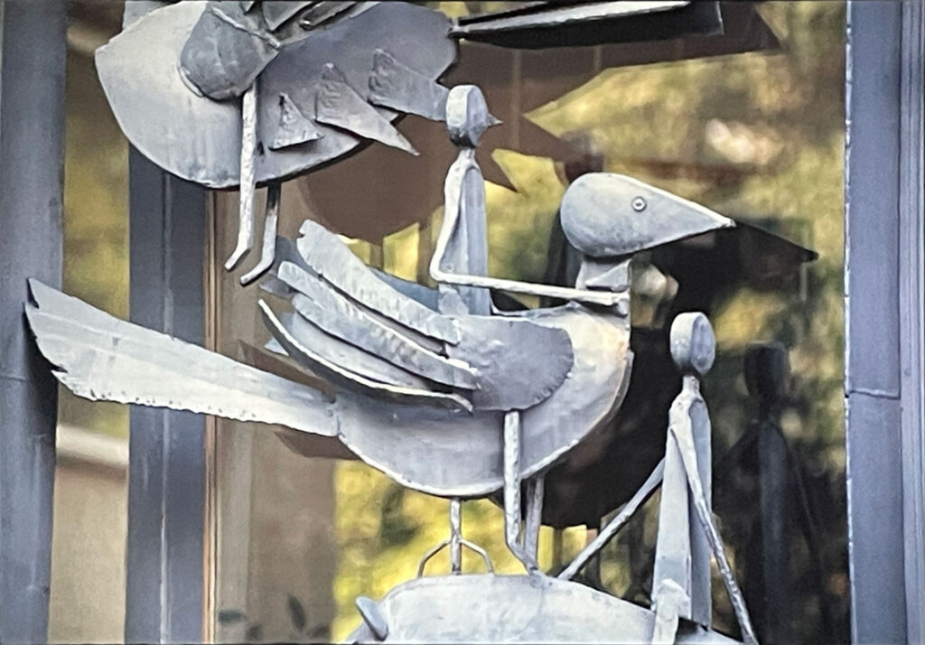 Close-up of a metal bird sculpture with layered wings and a perched figure beneath, attached to a building façade.