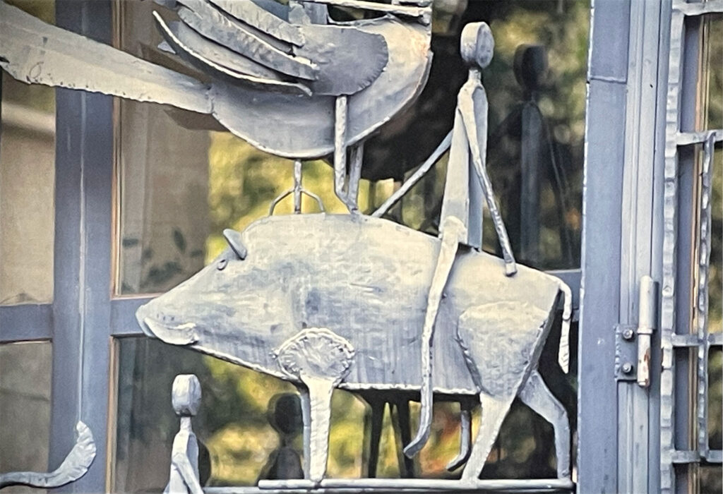 Detail of a metal sculpture showing a rider atop a boar, with simple elongated human forms against a reflective window.