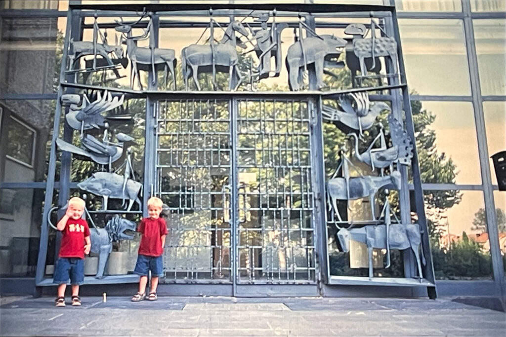 Two young children stand smiling before a large metal gate decorated with stylized animal and rider sculptures.