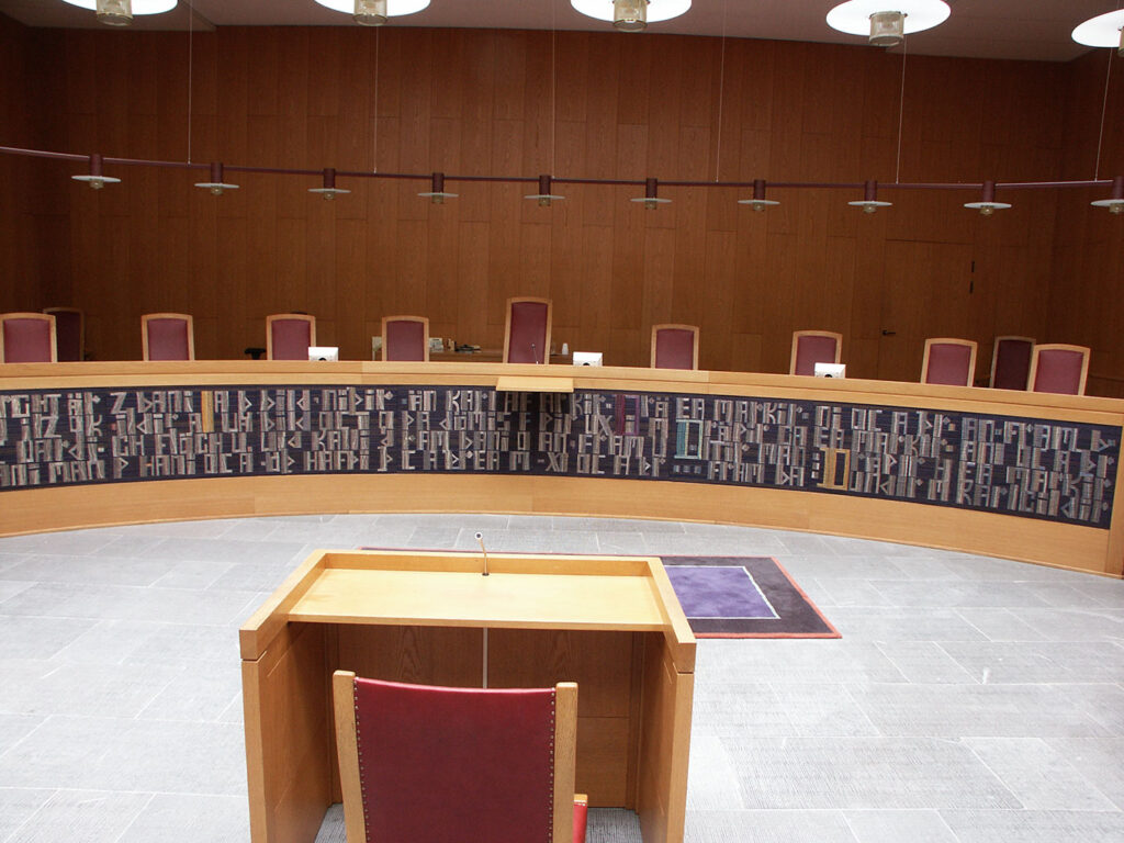 Wide courtroom interior with a curved bench lined by long woven panels of abstract lettering behind multiple seats.