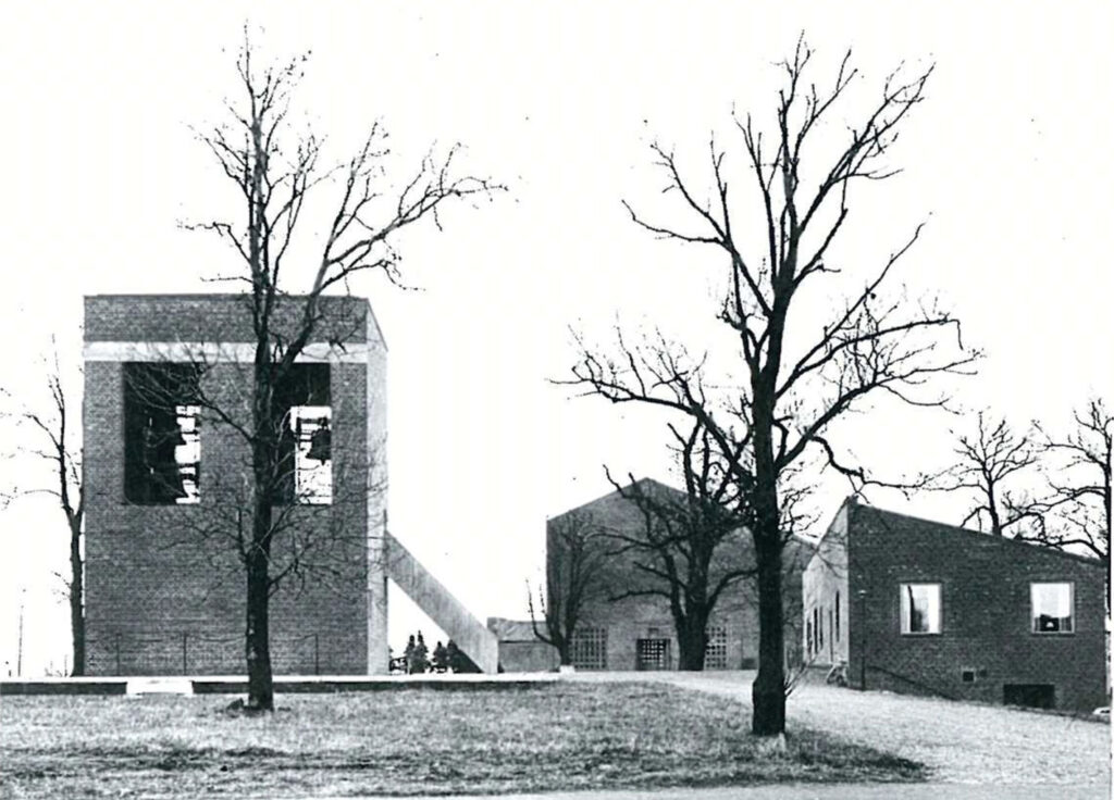 Black-and-white photo of a brick church complex with a tall bell tower, bare trees, and surrounding buildings on a grassy landscape.