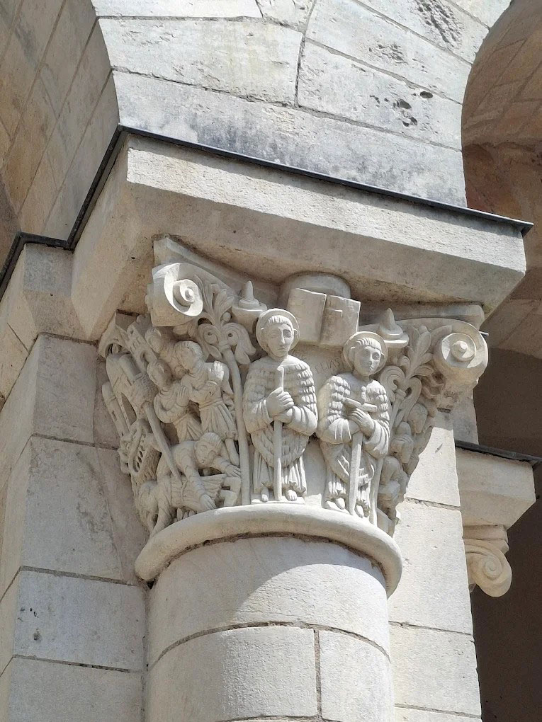 Stone capital featuring carved robed figures holding staffs amid intricate foliage and small scenes, set on a column of a historic building.