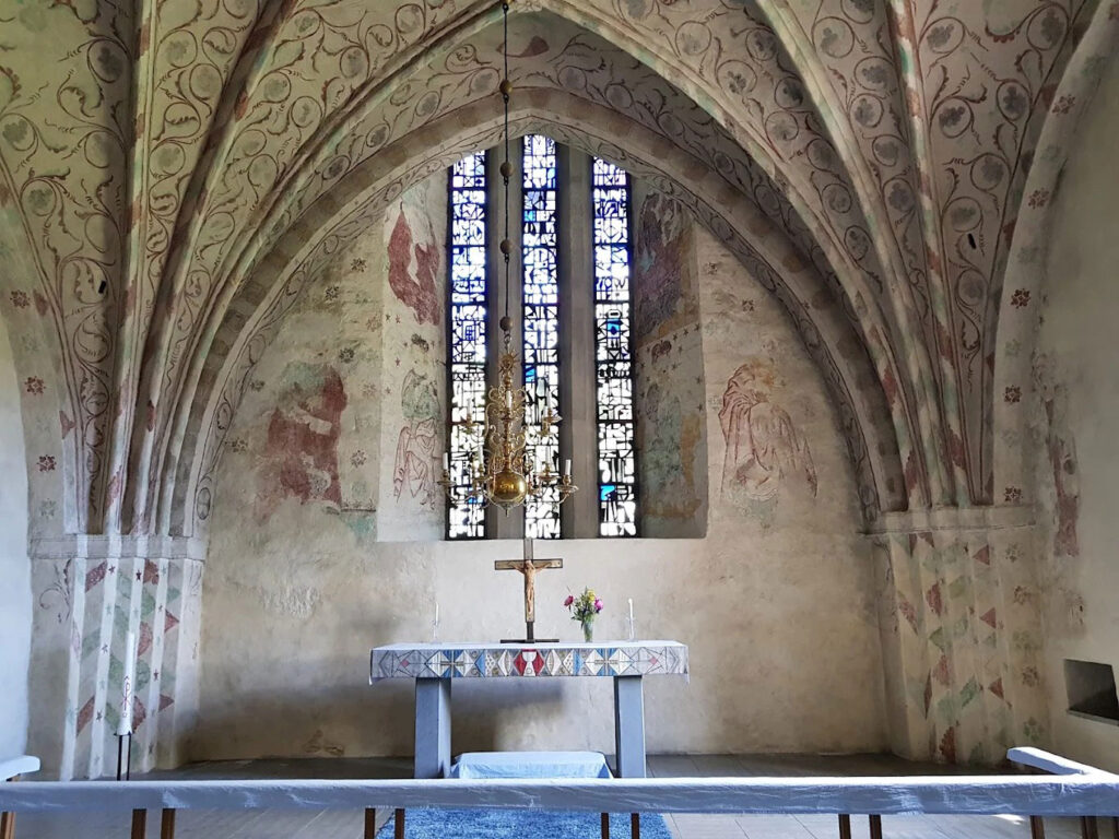 Gothic church interior with painted vaulted ceiling, faded wall murals, tall stained-glass window behind a simple altar and cross.