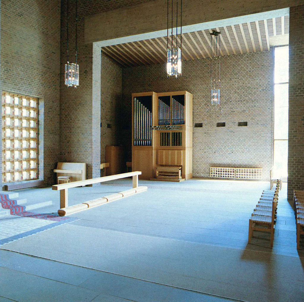 Modern church interior with brick walls, hanging lights, organ pipes, simple wooden benches, and patterned windows near the altar area.