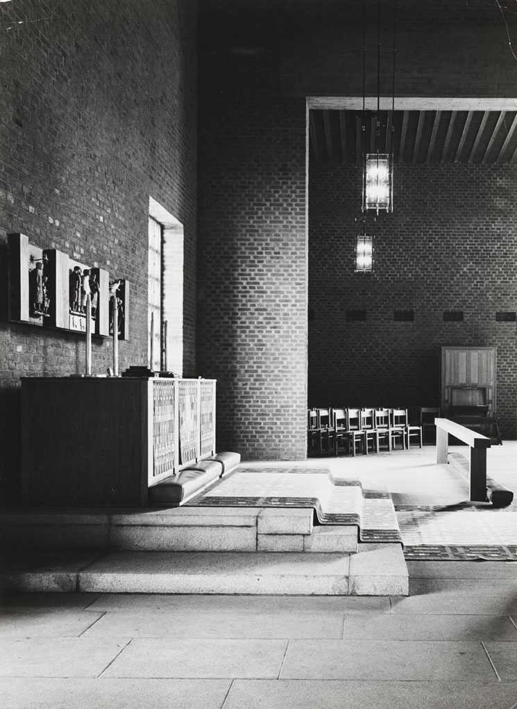 Black-and-white view of a minimalist brick church interior with stepped altar, hanging lamps, wooden furnishings, and rows of chairs.