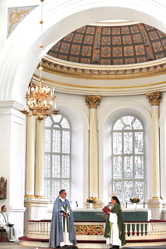 Clergy stand before an ornate church altar with tall arched windows, gold-trimmed columns, floral dome ceiling, and a glowing chandelier above.