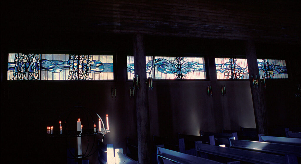 Stained-glass windows in a dim church interior glow with blue, teal, and purple figures, casting soft colored light over pews and candles.