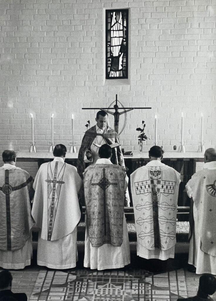 Black-and-white photo of priests in ornate vestments kneeling before an altar, with a celebrant reading beneath a narrow stained-glass window.