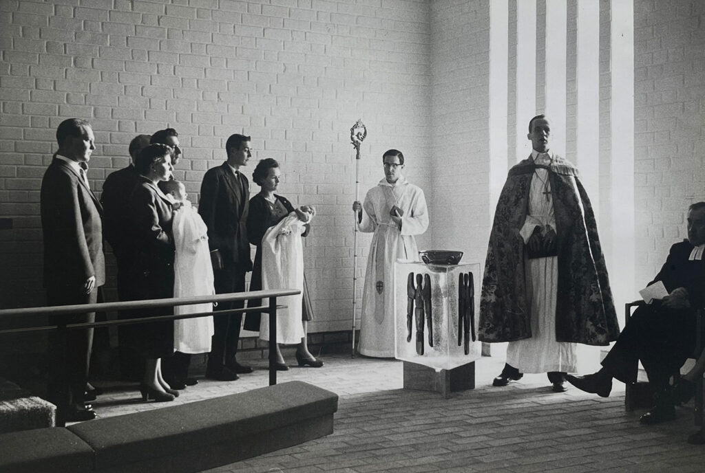 Black-and-white baptism scene with clergy beside a glass font and families holding infants, standing against a bright brick wall with tall windows.