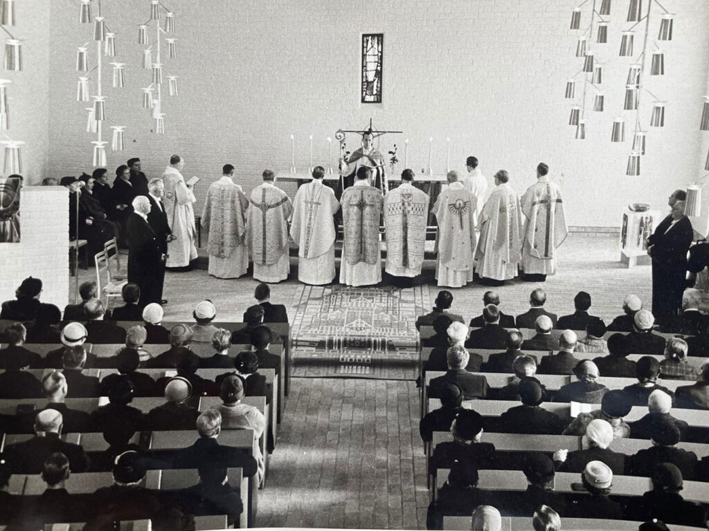 Black-and-white church service with robed clergy at the altar and a full congregation seated in pews beneath large hanging light fixtures.