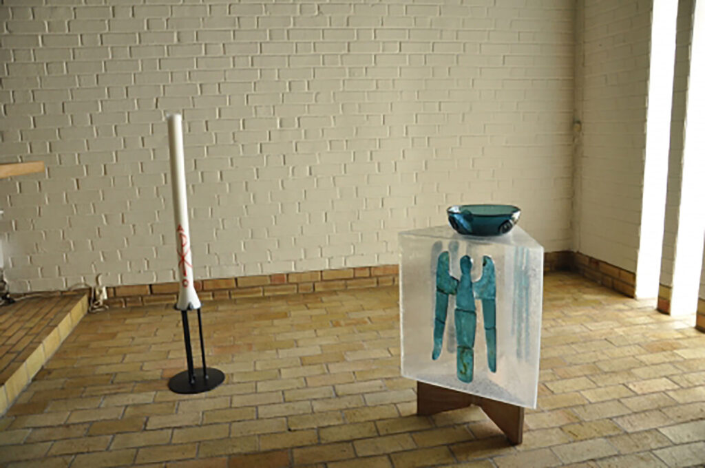 Baptismal area with a tall white candle and a translucent font featuring a blue angel design, topped by a blue glass bowl in a bright chapel.