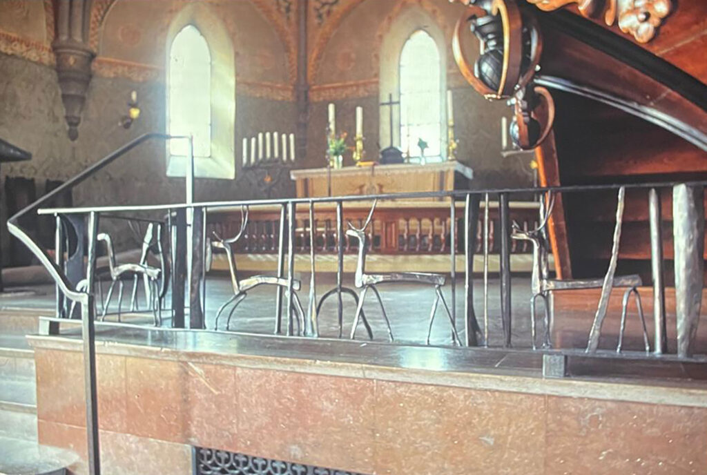 Metal church railing with stylized deer figures in front of an altar and tall arched windows, with warm light illuminating the interior.
