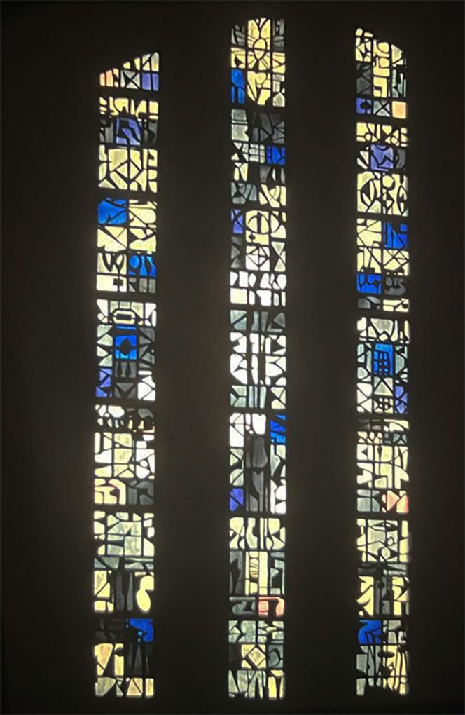 Three tall narrow stained-glass panels with abstract shapes in pale yellow, blue, and violet light glowing against a dark church interior.