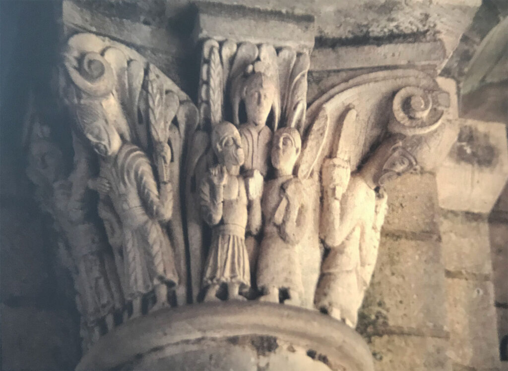 Stone capital carved with clustered standing figures in draped garments, framed by curled foliage and architectural details in soft light.