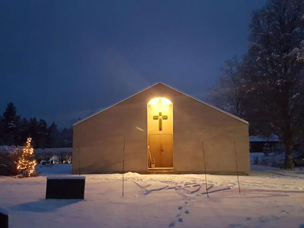 Small chapel in a snowy landscape at dusk, warmly lit above the entrance with a cross, footprints in the snow, and a lit tree nearby.