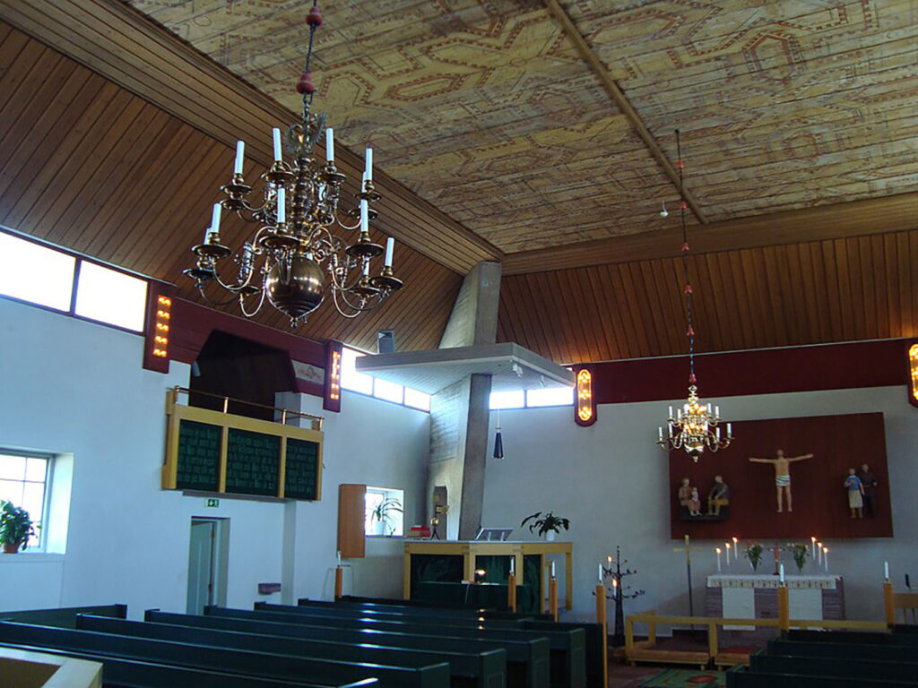 Interior of a church with green pews, chandeliers, patterned ceiling, wall art behind the altar, and soft daylight from high windows.