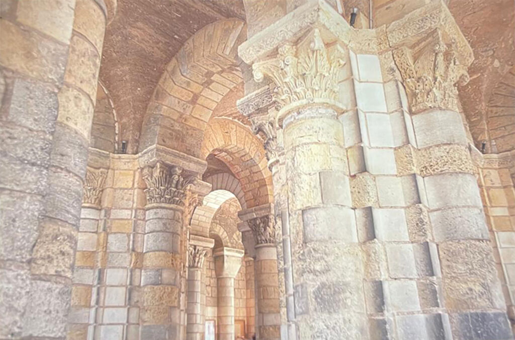 Interior view of a Romanesque church with massive stone columns, carved capitals, and rounded arches illuminated by soft, warm light.