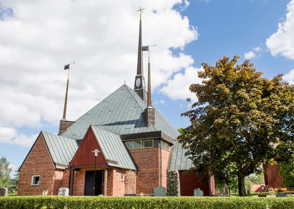 Exterior view of a brick church with steep metal roof, tall spires, small wooden entrance porch, trees and bright sky with clouds above.