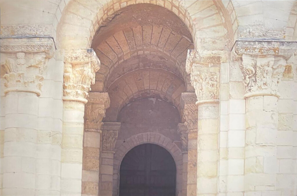 Romanesque church entrance with rounded stone arches, carved capitals featuring figures and foliage, and a dark wooden doorway beyond.