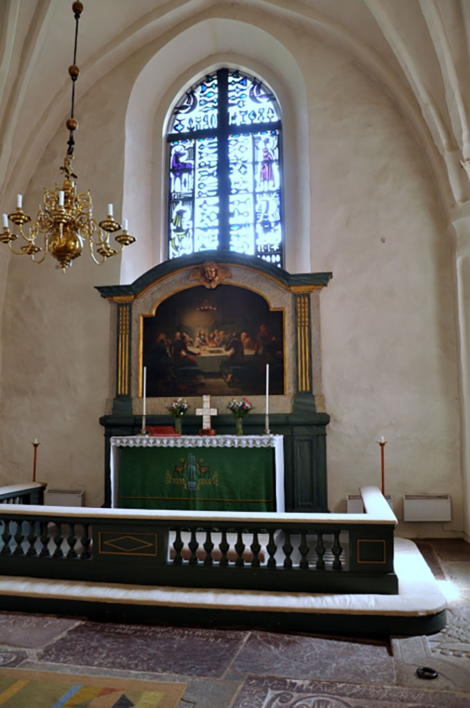 Church altar with green cloth, framed painting, brass chandelier, and tall stained-glass window illuminating the arched sanctuary interior.