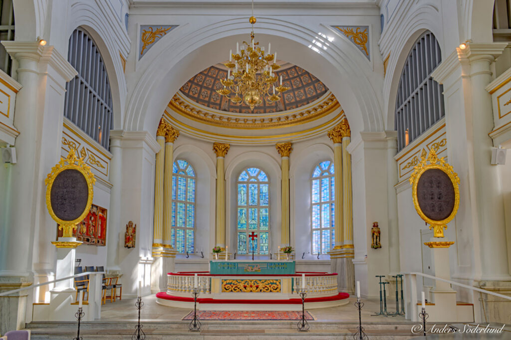 Bright church interior with ornate gold accents, arched ceilings, chandelier, tall stained-glass windows, and a decorated altar in soft light.